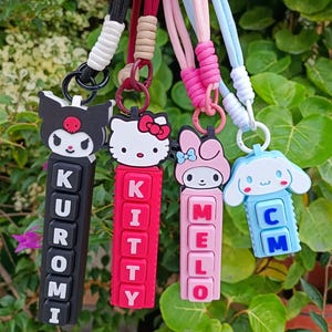 May include: Four Sanrio character keychains with lanyards. The black Kuromi keychain spells KUROMI, the red Hello Kitty keychain spells KITTY, the pink My Melody keychain spells MELO, and the light blue Cinnamoroll keychain spells CM.