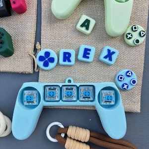 May include: A collection of gaming accessories, including a light blue controller with four buttons, and the letters "REX" in individual blue letters. Other items include a green controller, and a brown and beige lanyard.