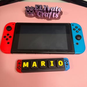 May include: A Nintendo Switch console with red and blue controllers, and a custom "MARIO" nameplate. The nameplate has red and blue ends, with yellow letters on a black background. The "ELYvate Crafts" logo is visible.