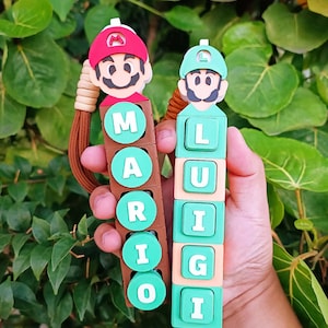 May include: Two keychains featuring cartoon characters Mario and Luigi. The Mario keychain has the name "MARIO" spelled out in green letters on brown blocks. The Luigi keychain has the name "LUIGI" spelled out in green letters on tan blocks.