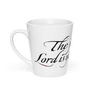 Może przedstawiać: Biały ceramiczny kubek do latte z zakrzywionym uchwytem. Kubek ma napis "The Lord is" w czarnej kaligrafii. Brązowy akcent znajduje się pod słowem "Lord".