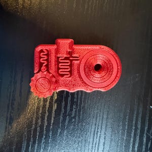 May include: A red 3D-printed object with a complex mechanical design. It features a circular gear, a series of curved lines, and a central hole. The object is likely a component or a model of a mechanical part.