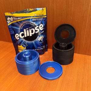 May include: A package of Eclipse Winterfrost gum next to three 3D-printed containers. Two containers are blue, one is black. The blue containers are cylindrical with a central post and a matching lid. The black container is also cylindrical with a lid.
