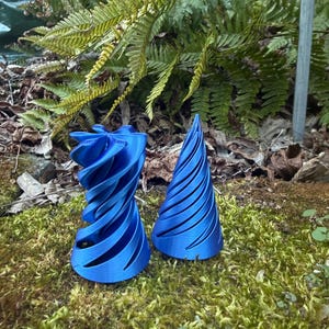 May include: Two blue, abstract, 3D-printed sculptures. One is a twisted, open form, and the other is a conical shape with spiral grooves. They are set on a bed of green moss, with ferns in the background.