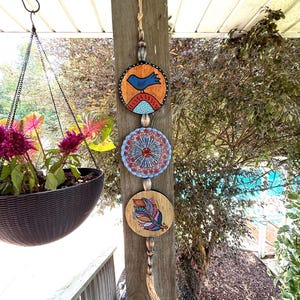 May include: A wooden wall hanging featuring three circular ornaments with painted designs. The top ornament has a blue bird, the middle one a floral pattern, and the bottom one a colorful feather. The ornaments are connected by a beaded string.