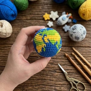 May include: A crocheted globe with blue oceans, green and yellow continents, held in a hand. Other crocheted items include a moon, stars, and a rocket. Yarn balls in blue, green, and yellow are in the background. Gold scissors and crochet hooks are also present.