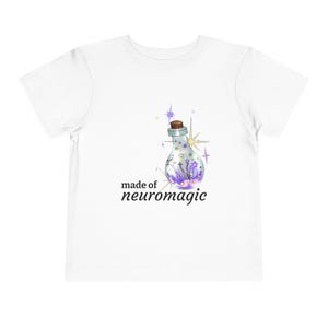 May include: White toddler t-shirt featuring a graphic of a glass bottle with a cork stopper, filled with purple and yellow stars and sparkles. The text "made of neuromagic" is printed below the bottle in black.