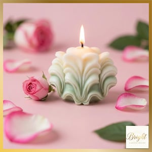 Op de afbeelding: Een brandende, wit en lichtgroene bloemkaars. De kaars is omgeven door roze rozenblaadjes en een kleine roze roos op een roze ondergrond. De kaars heeft een decoratief ontwerp en maakt deel uit van een home decor thema.
