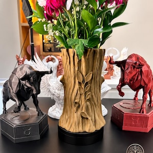 Könnte beinhalten: Eine beigefarbene Baumstumpf-Vase mit einem Ast- und Blattrelief, flankiert von einer dunkelgrauen geometrischen Stierstatue links und einer roten rechts, jeweils auf einem sechseckigen Sockel mit dem Stier-Sternzeichen-Symbol.