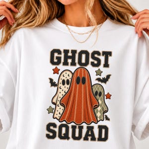 Könnte beinhalten: Ein weißes Sweatshirt mit Rundhalsausschnitt und einem Grafikdruck mit dem Text GHOST SQUAD in schwarzen Blockbuchstaben. Das Design zeigt drei stilisierte Geister in Orange, Creme und Olivgrün mit Mustern, umgeben von kleinen Fledermäusen und Sternen.