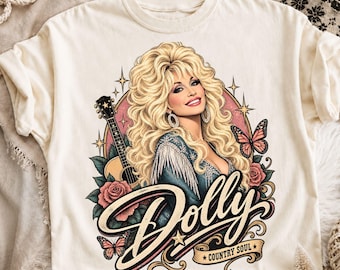 Può includere: Una t-shirt color crema con un'illustrazione grafica di una donna bionda con capelli voluminosi, una chitarra, rose e farfalle. Il testo "Dolly" è scritto in grande corsivo, con un banner sottostante che recita "COUNTRY SOUL" e delle stelle.