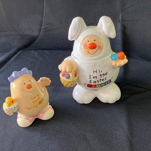 Puede incluir: Dos figuras de cerámica de Pascua. Una es un conejito blanco con el texto "Hi, I'm the Easter Beanie". La otra es una figura de color canela con el texto "I'm a REAL Spring". Ambas figuras tienen detalles coloridos y están sobre un fondo oscuro.