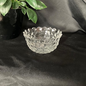 Puede incluir: Un cuenco pequeño de vidrio transparente con un patrón geométrico. El cuenco tiene un borde festoneado y probablemente se utiliza para servir pequeñas porciones de comida o como objeto decorativo. El cuenco está sobre una superficie negra.