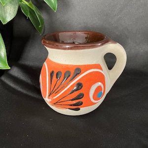 Op de afbeelding: Kleine, handbeschilderde keramische kruik met een afgeronde body en een handvat. De kruik heeft een crèmekleurige basis met oranje en zwarte bloemmotieven. De rand en de binnenkant zijn bruin. Een klein, decoratief stuk.