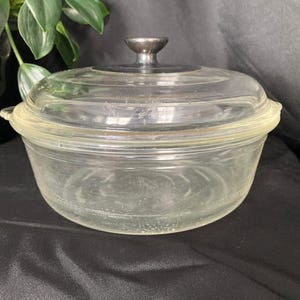 May include: A clear glass casserole dish with a matching lid. The dish has a rounded shape with a slightly flared rim and two small handles. The lid has a silver-coloured knob. The dish is on a black surface.