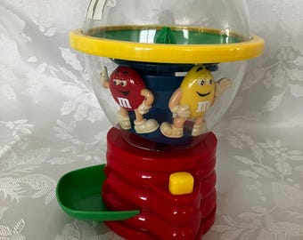 May include: A plastic M&M's candy dispenser featuring a clear spherical top with a green lid, a yellow ring, and a red base. Inside the clear globe are red and yellow M&M character figures. A small yellow button sits on the red base next to a green tray.