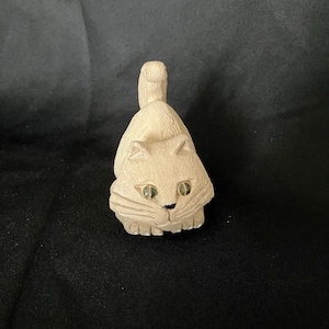May include: A small, light beige carved wooden cat figurine with a textured surface, pointed ears, and two inset green glass eyes. The cat is depicted in a seated position with its tail curled upward, featuring carved whiskers and small paws.