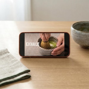 Pode incluir: Um smartphone exibe a palavra "EXHALE" durante a prepara&ccedil;&atilde;o do ch&aacute; matcha. Um batedor de bambu mistura ch&aacute; verde numa tigela de cer&acirc;mica. Outra tigela de cer&acirc;mica e um guardanapo de linho dobrado est&atilde;o sobre a superf&iacute;cie de madeira.
