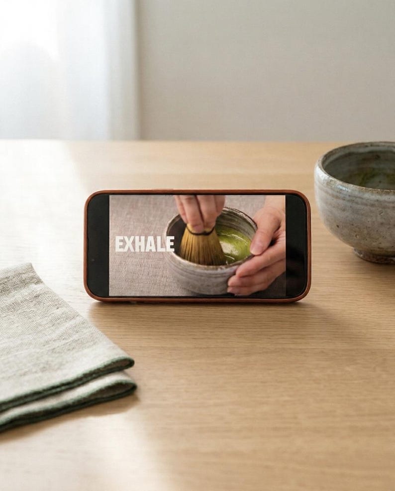 Pode incluir: Um smartphone exibe a palavra "EXHALE" durante a prepara&ccedil;&atilde;o do ch&aacute; matcha. Um batedor de bambu mistura ch&aacute; verde numa tigela de cer&acirc;mica. Outra tigela de cer&acirc;mica e um guardanapo de linho dobrado est&atilde;o sobre a superf&iacute;cie de madeira.