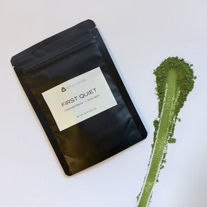 Pode incluir: Uma embalagem preta de "FIRST QUIET Ceremonial Matcha Kyoto Japan" com uma etiqueta branca, ao lado de uma faixa de p&oacute; de matcha verde sobre uma superf&iacute;cie branca. A embalagem est&aacute; rotulada "NET WT. 30 g (1.06 OZ)".