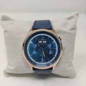 May include: A luxury wristwatch with a blue face and a rose gold case. The watch features a blue leather band and is displayed on a white pillow. The watch face displays the date and the brand name "Jaeger-LeCoultre".