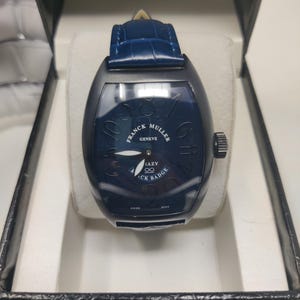 May include: A Franck Muller Geneve Crazy Black Badge watch with a tonneau-shaped black case and a dark blue dial featuring large, non-sequential numerals. The timepiece includes a blue leather strap with a crocodile texture and a black crown.