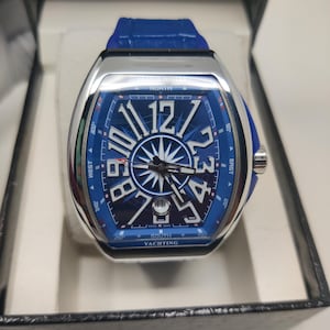 Könnte beinhalten: Eine Franck Muller Vanguard Yachting Uhr mit silberfarbenem Tonneau-Gehäuse und blauem Lederarmband. Das blaue Zifferblatt zeigt große weiße arabische Ziffern, ein zentrales Kompassrosen-Design, ein Datumsfenster bei sechs Uhr und Himmelsrichtungen.