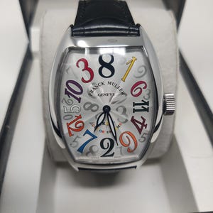 May include: A Franck Muller Geneve wristwatch with a tonneau-shaped silver case and a black leather strap. The white dial features a guilloche pattern and large, multi-colored Arabic numerals. The text reads Franck Muller Geneve Color Dreams Swiss Made.