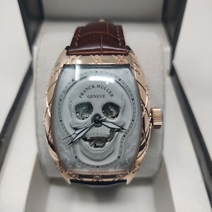 May include: A Franck Muller Geneve wristwatch with a rose gold tonneau-shaped case featuring a white skull dial, skeletonised eye and mouth openings, black hands, and a brown leather strap with a textured pattern, presented in a black box.