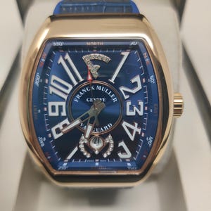 Puede incluir: Un reloj Franck Muller Vanguard con caja tonneau en tono oro rosa y esfera azul. La esfera presenta grandes números arábigos blancos, una ventana de fecha con detalles de diamantes y un logotipo F con diamantes. Correa de cuero azul.