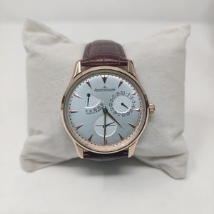 May include: A luxury wristwatch with a light blue face, gold-tone case, and brown leather strap. The watch features multiple sub-dials and the Jaeger-LeCoultre logo. The watch is displayed on a white cushion.