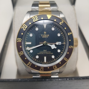 May include: A Tudor Geneve GMT Chronometer watch with a black dial, gold-tone hands, and a two-tone stainless steel and gold bracelet. The bezel features a black and burgundy insert with gold numerals, and the dial displays the text Tudor Geneve GMT.