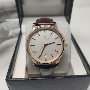 May include: A luxury wristwatch with a brown leather strap and a gold-tone case. The watch face is white with gold-tone hands and hour markers. The watch is displayed in a black presentation box.