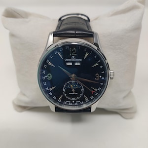 May include: A luxury wristwatch with a dark blue face, silver casing, and a black leather band. The watch displays the date, month, and moon phase. The watch is resting on a white pillow.