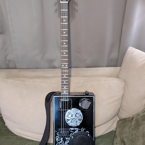 Könnte beinhalten: Eine schwarze Zigarrenkistengitarre mit einem Holz Hals und Griffbrett. Auf der Kopfplatte steht "BUSKER" in weißer Schrift. Der Korpus hat einen Lautsprecher, eine Grafik eines Gesichts und den Text "THE BIG..." und "Voodoo Child". Ein schwarzer Gurt ist befestigt.