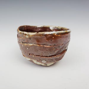 May include: A handmade ceramic bowl with a textured, layered design. The bowl features a rich, earthy brown color with lighter, speckled accents and a white rim. The unique shape and finish suggest a rustic, artisanal aesthetic.