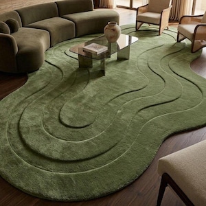 May include: A large, olive green area rug featuring a distinct, organic, wavy, layered design with raised, curved sections that create a textured, topographical appearance across the floor, covering a significant portion of the room's wooden surface.