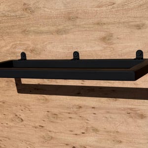 Könnte beinhalten: Ein schwarzes, rechteckiges schwebendes Regal mit minimalistischem Design. Das Regal ist an einer Holzwand befestigt und zeichnet sich durch eine schlichte, klare Ästhetik aus. Das Regal wird von zwei Halterungen getragen.