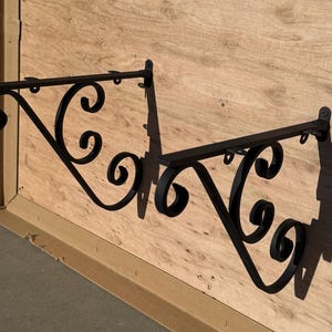 May include: Two black wrought iron sign brackets with decorative scrollwork. The brackets are mounted on a light brown wooden surface. Each bracket has a horizontal bar with small hooks and a curved, ornate design.