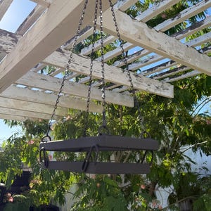 May include: A hanging, two-tiered metal planter with a dark gray finish, suspended by chains from a white pergola. The planter has a rectangular shape with curved handles. Lush green foliage and a blue sky are in the background.