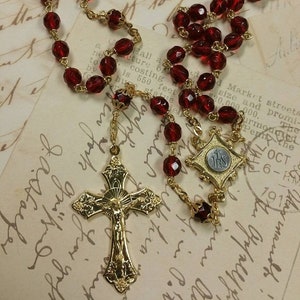 Puede incluir: Un rosario de vidrio rojo con detalles dorados. El rosario presenta un crucifijo dorado con una figura de Jesucristo y una medalla dorada con la inscripción "IHS".