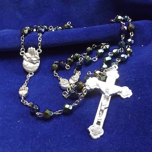 Puede incluir: Un rosario de plata con cuentas negras y un crucifijo de plata. Las cuentas son iridiscentes y tienen un tono verde oscuro. El crucifijo presenta una figura de Jesucristo.