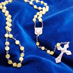 May include: A yellow glass rosary with a silver cross and a silver charm with a circular design. The rosary is on a blue velvet background.