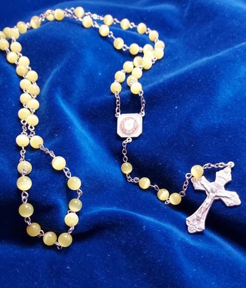 May include: A yellow glass rosary with a silver cross and a silver charm with a circular design. The rosary is on a blue velvet background.