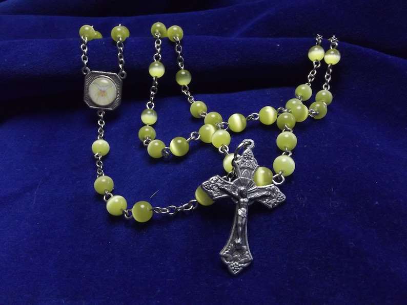 May include: A silver rosary with a silver crucifix and a round, white, and silver pendant. The beads are light green and have a pearlescent finish.