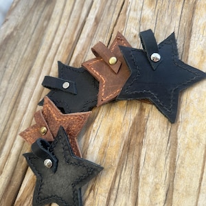 May include: Star-shaped leather keyrings in shades of brown and black. Each keyring has a stitched border and a small strap with a metal rivet. The keyrings are displayed on a wooden surface.
