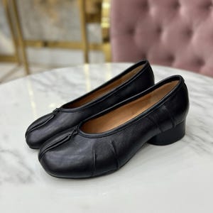 May include: A pair of black leather shoes with a low heel. The shoes have a rounded toe and a small bow detail on the top. The shoes are sitting on a white marble surface.