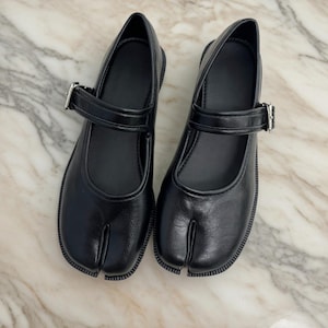 May include: Pair of black leather Tabi shoes. These split-toe shoes have a Mary Jane style with a strap and buckle closure. The soles are thick and have a textured edge.