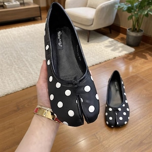 May include: Black split-toe ballet flats with white polka dots. The shoes have a rounded toe and a small bow detail. One shoe is held in a hand, and another is on the floor. The shoes are a size 36.