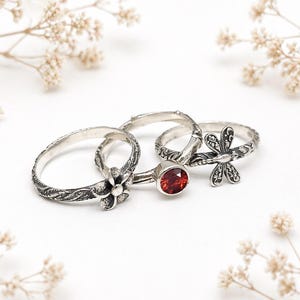 May include: Four silver rings with detailed designs are presented. One ring has a flower, another a red gemstone, and a third a dragonfly. The rings feature floral and textured patterns, creating a delicate and artistic appearance. The rings are displayed on a white surface.